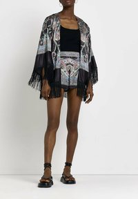 Zwarte kimono met een ingewikkeld paisleypatroon in roze en blauw, met franjeranden, gedragen over een zwart tanktop en shorts.