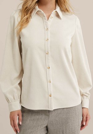 Vrouw draagt een crèmekleurige geribde blouse met knopen, lange mouwen met opgepofte manchetten en grijze broek met visgraatpatroon.