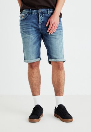 Mand iført opkaldte blå denimshorts, hvide crew-strømper, sorte sneakers med brune såler, stående mod en hvid baggrund.