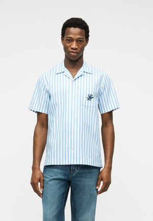 Mann trägt ein kurzärmeliges, blau-weiß gestreiftes Hemd mit Knöpfen und blaue Jeans, steht vor einem schlichten Hintergrund.