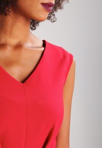 Robe rouge sans manches avec un décolleté en V, présentant une texture lisse et un détail de pli subtil au centre.