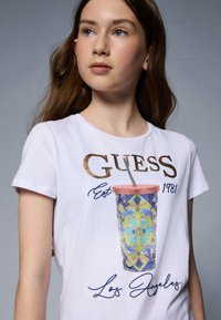 Camiseta blanca con la frase "GUESS Est 1981" en letras doradas. Gráfico de un vaso decorado colorido, con intrincados detalles de cuentas y una pajita.