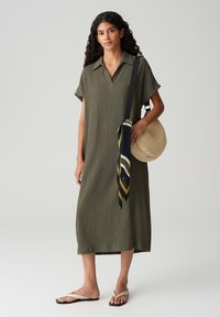 Olive green midi dress with short sleeves, collar, and vertical ribbed texture. Paired with a woven circular bag and a patterned scarf.