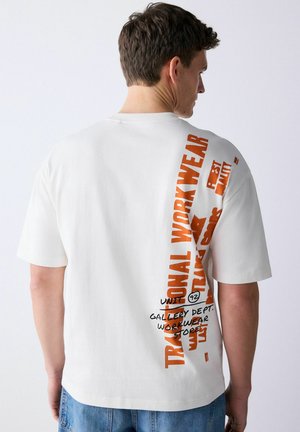 Junger Mann trägt ein weißes T-Shirt mit kurzen Ärmeln und vertikalem Text in Orange und Schwarz auf dem Rücken, kombiniert mit blauen Jeans.