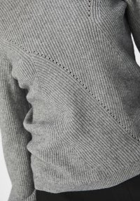 Záclose-up šedého ribovaného pleteného svetru s diagonálními texturovanými vzory a malými detaily v středu. 