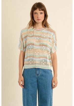 Giovane donna che indossa un top corto a maniche corte lavorato a maglia multicolore pastello e jeans blu a vita alta con gamba larga, in piedi contro uno sfondo semplice.