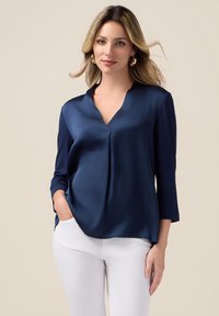 Navy satin blouse with a V-neck, three-quarter sleeves, and a relaxed fit. Features a subtle pleat down the center, paired with white pants.