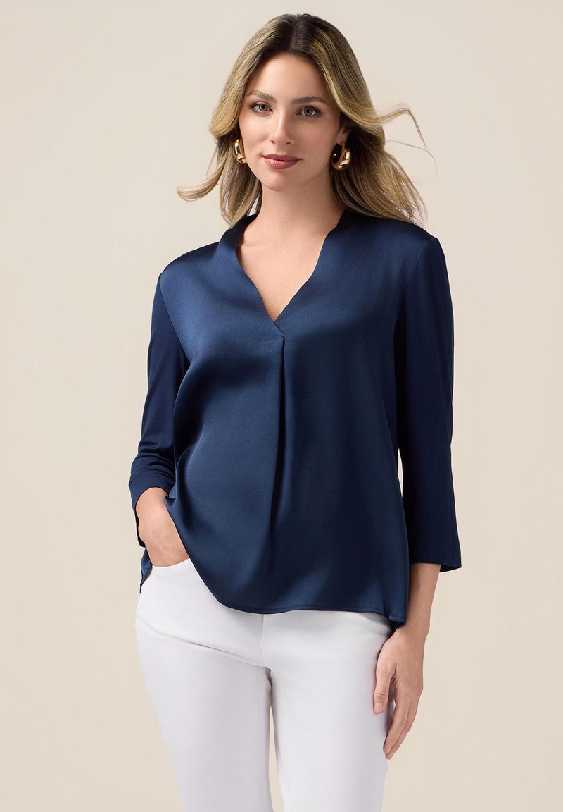 Navy satin blouse with a V-neck, three-quarter sleeves, and a relaxed fit. Features a subtle pleat down the center, paired with white pants.