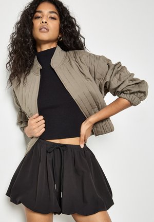 Vrouw met krullend haar die een cropped zwart topje draagt, een olijfgroene gewatteerde jas met geplooide mouwen en zwarte shorts met trekkoord, poserend met handen in de taille.