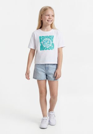 Chica sonriente con cabello largo y rubio, vistiendo una camiseta blanca con la frase "Friday Mood", shorts de mezclilla claros y zapatillas blancas, de pie frente a un fondo blanco.