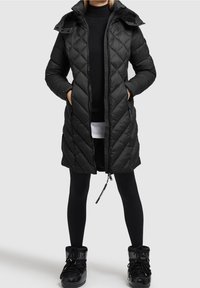 Parka acolchada negra con capucha, mangas largas y cremallera en la parte delantera. Cuenta con bolsillos laterales y una tela texturizada. Usada sobre leggings negros y botas.