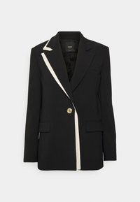 Blazer noir ajusté avec bordure blanche sur un revers et une fermeture à un bouton doré, présenté sur un fond blanc.