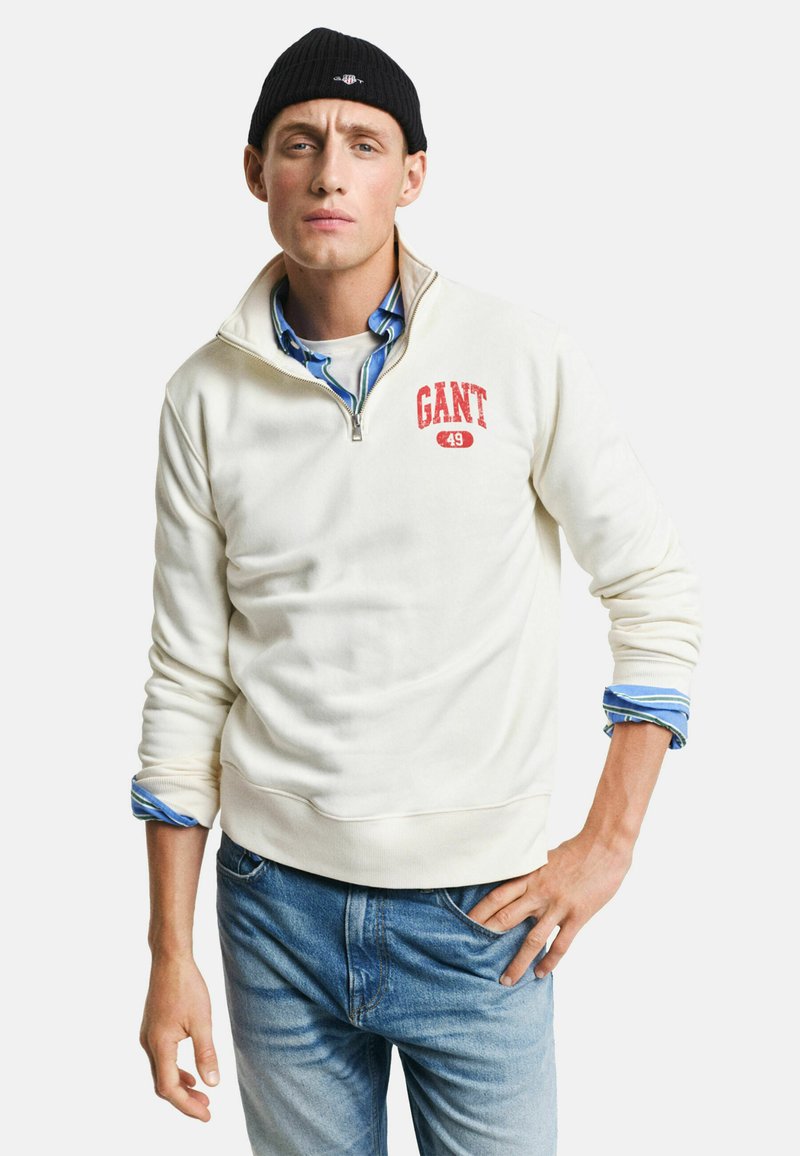 Witte fleece sweatshirt met een kwart rits, voorzien van een rood "GANT 49" logo. Het is gedragen over een blauw geruit shirt, gecombineerd met blauwe denim jeans.