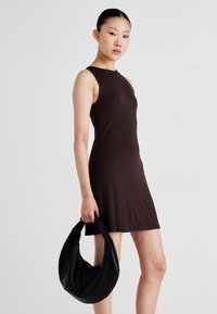 Bruine mouwloze jurk van zacht materiaal, met een getailleerde silhouet en een gebogen halslijn. Model draagt een soepele zwarte hobo tas.