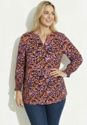Uśmiechnięta kobieta o blond włosach ubrana w bluzkę z długim rękawem z pomarańczowo-fioletowym kwiatowym wzorem i ciemnoniebieskie jeansy, z ręką na biodrze.