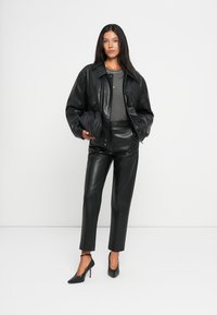 Veste oversize en cuir noir associée à un haut gris ajusté et un pantalon en cuir noir. Le mannequin porte des chaussures à talons hauts noires et tient un sac.