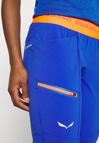 Siniset ulkoiluhousut, joissa on oranssi vyötärö, sivutasku oranssilla vetoketjulla ja logotiedot. Kangas on sileä ja kestävä.