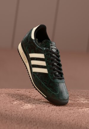 Green textured sneaker with a snake-skin pattern, featuring three cream stripes, black laces, and a black outsole. Classic Adidas branding.