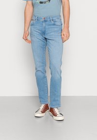 Homem com jeans azul claros e ténis vermelhos de cano alto, de pé contra um fundo claro e liso.