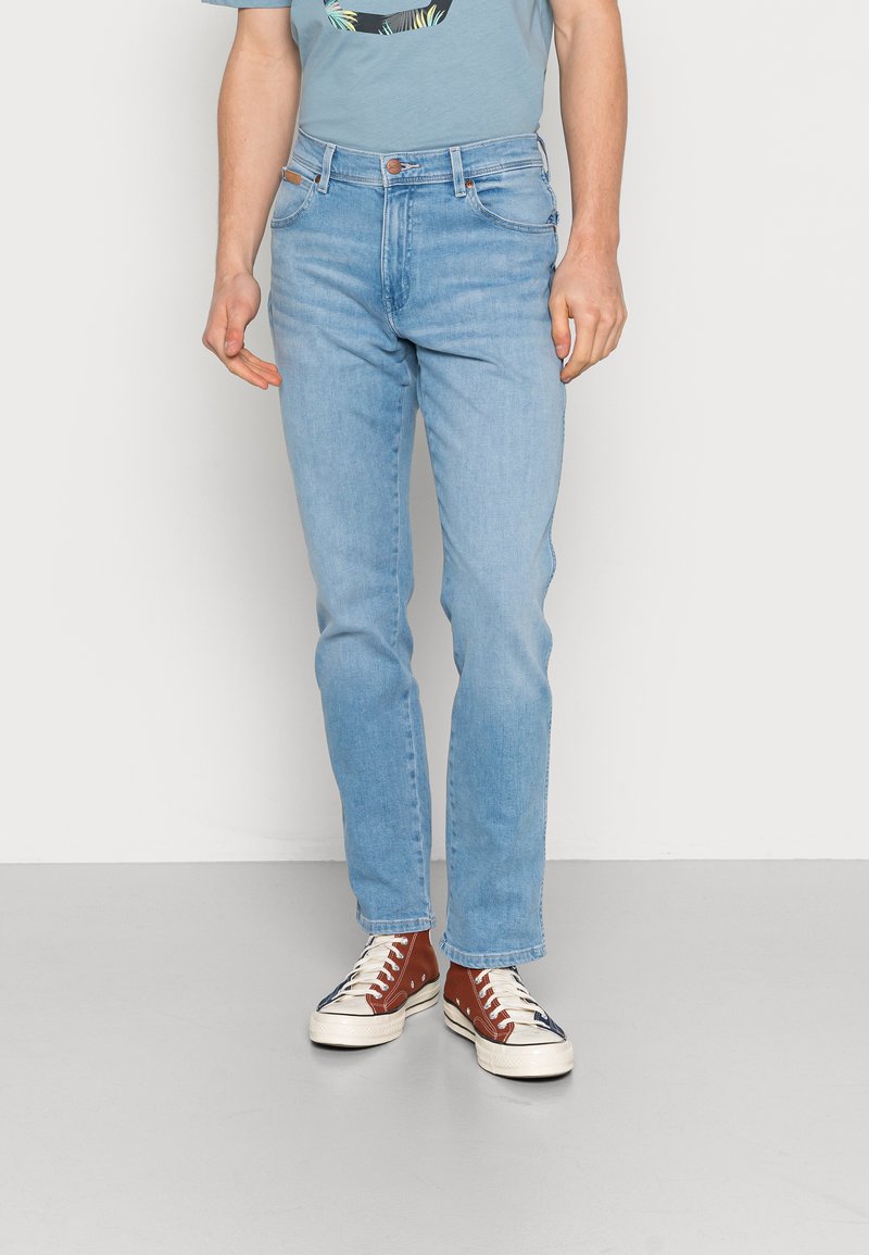 Homem com jeans azul claros e ténis vermelhos de cano alto, de pé contra um fundo claro e liso.