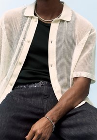 Górna część ciała osoby noszącej kremową siateczkową koszulę na guziki, czarny top na ramiączkach, pasek z gwiazdkami, naszyjnik z pereł oraz srebrną bransoletkę.