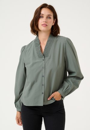 Groene blouse met kraag en geplooide halslijn, lange poffende mouwen en knopen aan de voorkant. De stof lijkt soepel en lichtgewicht te zijn.