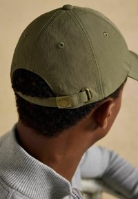 Mann mit olivgrüner Baseballmütze mit verstellbarem Riemen und Messingschnalle, grauem, geripptem Pullover, von hinten und leicht seitlich gesehen.