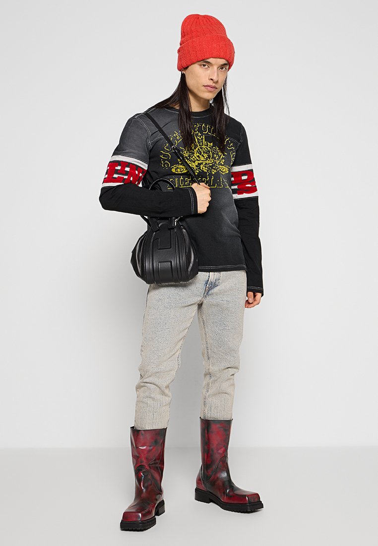 Schwarzes Langarmshirt mit gelbem Grafikdruck, roten Akzenten; helle Jeans; glänzende rot-schwarze Stiefel bis zur Wade; schwarze Handtasche. Rote Strickmütze.