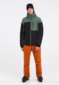 Gepolsterde zwarte en groene jas met een ritsborstzak, in combinatie met oranje broek en zwarte skiboots. Model draagt een zwarte beanie.