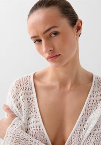 Cardigan alb tricotat cu deschidere în față, având un model din dantelă și o croială lejeră. Textura este ușoară și aerată, ideală pentru a fi purtată în straturi.