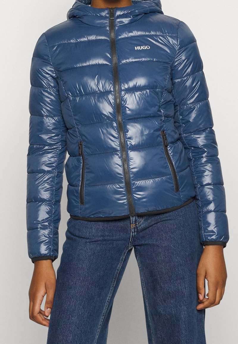 Marineblaue Steppjacke mit glänzender Oberfläche, ausgestattet mit einem Reißverschluss vorne, Kapuze und seitlichen Taschen. Mit einem Logo auf der Brust und einem gerippten Saum.