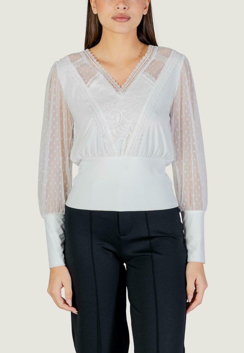 Biała bluzka z przejrzystymi rękawami w groszki, koronkowymi zdobieniami na gorsecie i dopasowaną talią. W zestawieniu z czarnymi spodniami z wysokim stanem.