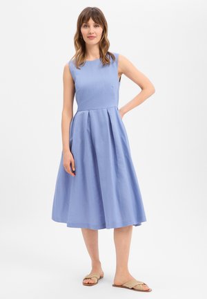 Vrouw staand in een mouwloze, lichtblauwe midijurk met plooien, beige platte sandalen dragend tegen een effen witte achtergrond.