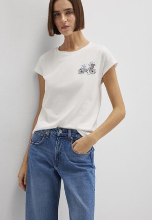 Frau mit kurzen braunen Haaren, die ein weißes T-Shirt mit kleinem blauem Fahrradmotiv und blaue Jeans trägt, steht vor einer weißen Wand.