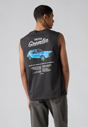 Homme portant un débardeur noir avec un dessin de voiture vintage bleue et le texte « Denim Gremlin » dans le dos, associé à un jean gris foncé.