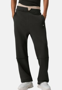 Schwarze Jogginghose aus glattem Stoff, mit beigefarbenem elastischem Bund, Seitentaschen und einem kleinen Logodetail an einem Bein.