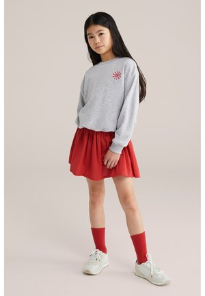 Dekle z dolgimi temnimi lasmi, oblečeno v sivo majico s sončnim rdečim motivom, rdečo krilo, rdeče nogavice in bele superge, stoji pred nevtralnim ozadjem.