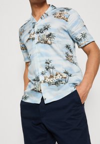 Chemise à manches courtes avec col en tissu bleu clair, présentant un motif tropical avec des palmiers et des scènes de plage, agrémenté d'accents floraux blancs.