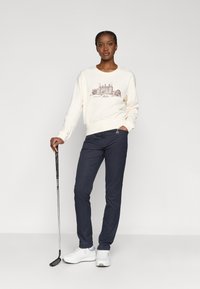 Sudadera crema con un estampado arquitectónico marrón, combinada con pantalones azul marino. El modelo sostiene un palo de golf y lleva zapatillas blancas.