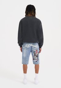Felpa grigia a maniche lunghe, abbinata a pantaloni shorts in denim blu con un teschio ricamato colorato e scritte. Sneakers bianche completano l'outfit.