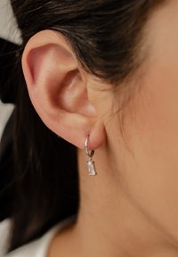 Pendiente de aro de plata con una gema rectangular y transparente colgando del aro. El metal suave y la piedra transparente contrastan de manera elegante.