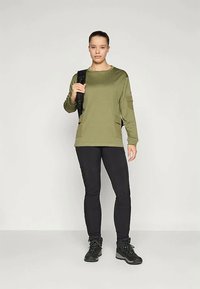 Olajzöld, hosszú ujjú felső oldalzsebekkel, fekete, testhez simuló nadrággal és fekete túracipővel párosítva, semleges háttér előtt.