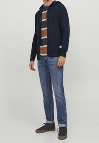 Námořnická mikina na zip s podšívkou kapuce, kombinovaná se pruhovaným tričkem v zemitých odstínech, modrými džíny a tmavými teniskami s hnědými podrážkami.