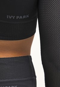 Närbild av en person som bär svart Ivy Park träningskläder med andningsbar mesh-tyg vid armen, midjan och överkant.