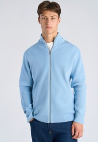 Giovane uomo con capelli castani corti che indossa un maglione a costine azzurro con zip e jeans scuri, in piedi davanti a uno sfondo chiaro e semplice.