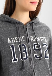Sudadera gris con cremallera y cordones, que presenta la inscripción en blanco y marino "ABERC ROMBIE 1892" en el frente, confeccionada en un tejido suave y texturizado.