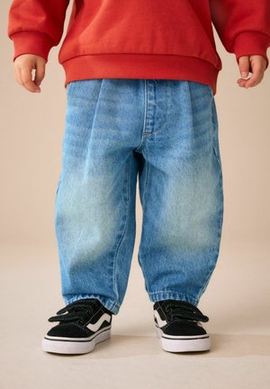 Dziecko ubrane w luźne jasnoniebieskie jeansy, czarno-białe sneakersy i czerwoną bluzkę z długim rękawem, stojące na jasnej podłodze na tle beżowej ściany.