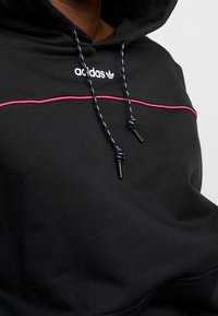 Černá mikina s kapucí s růžovým horizontálním pruhem a bílým logem adidas. Má vzorované šňůrky a jemnou texturu. Mikina je s kapucí.