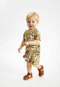 Kortärmad outfit och shorts med ett gult banantryck på vit bakgrund, parat med bruna sandaler och hållande bruna solglasögon.