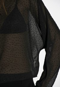 Durchsichtige schwarze Bluse mit funkelnden schwarzen Verzierungen, weiten Ärmeln und einer enganliegenden schwarzen Unterlage. Die Textur wirkt leicht und luftig.